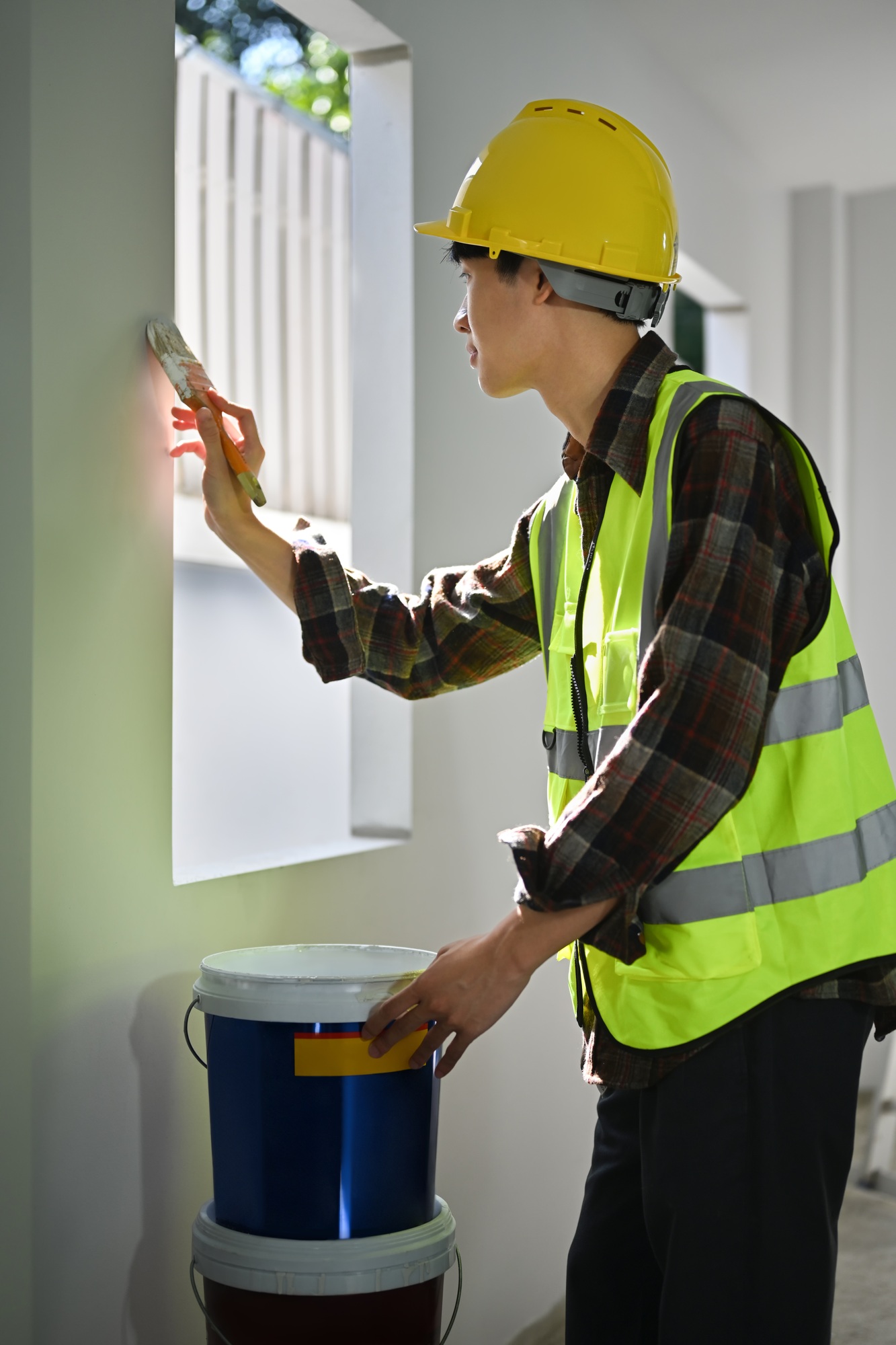 Man painter in safety helmet and yellow vest holding painting brush painting a white wall.